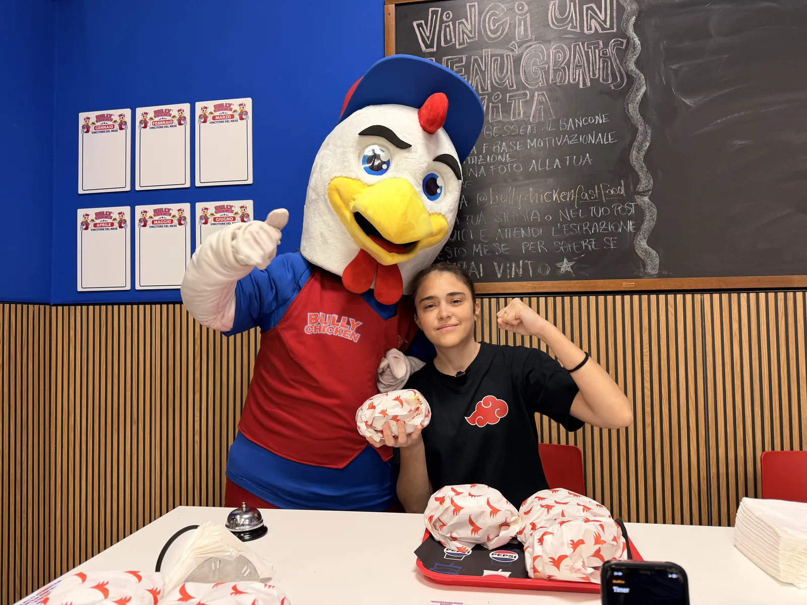 Chiara della sfida Chiara contro tutti seduta al tavolo di Bully Chicken mentre flette il bicipite con un panino incartato in mano, accanto alla mascotte del locale, davanti al vassoio con gli altri panini