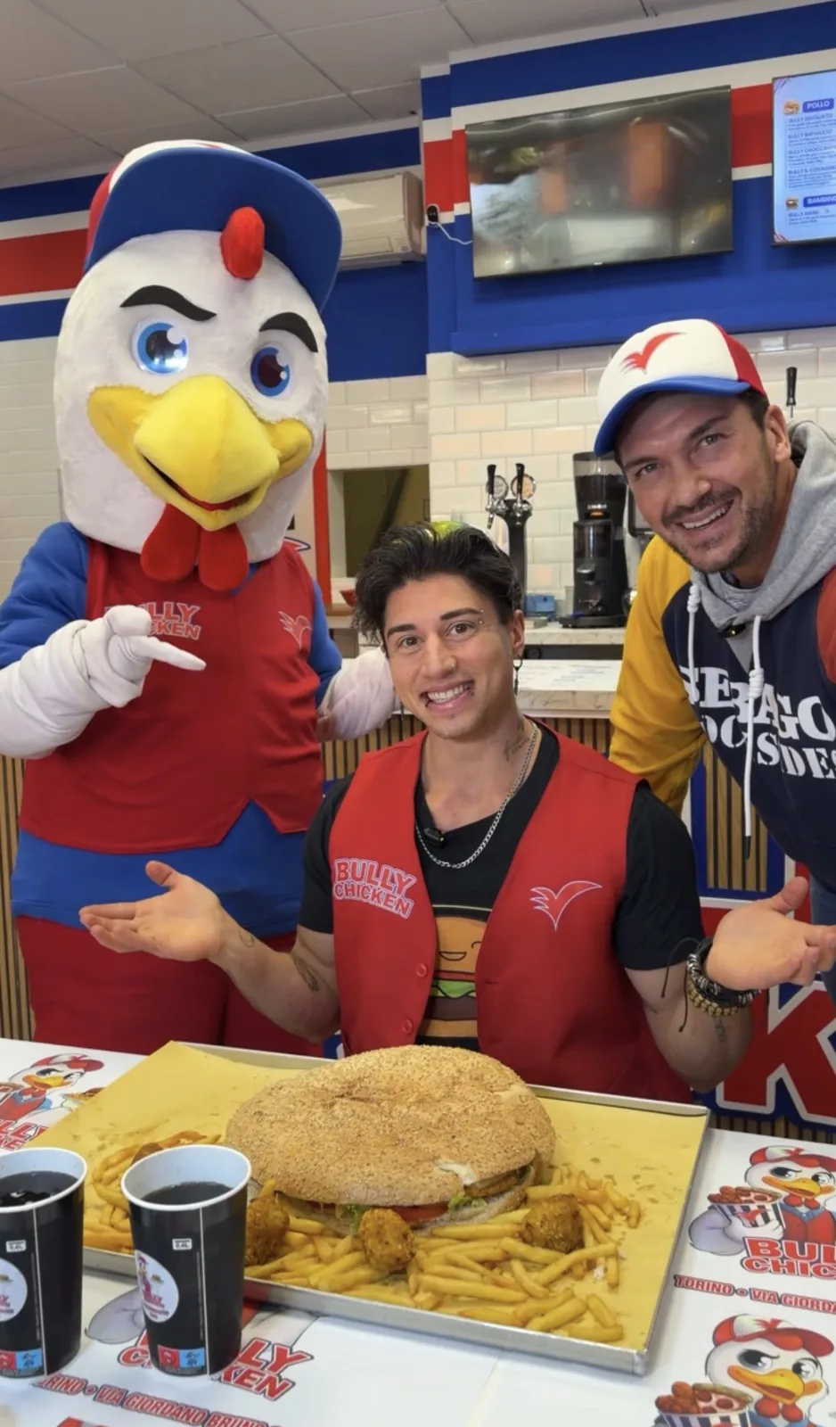 Paki con il gilet rosso di Bully Chicken seduto davanti al panino da 2,5 kg dell'XL Bully Challenge, con la mascotte a sinistra e Stefano di Bully Chicken a destra